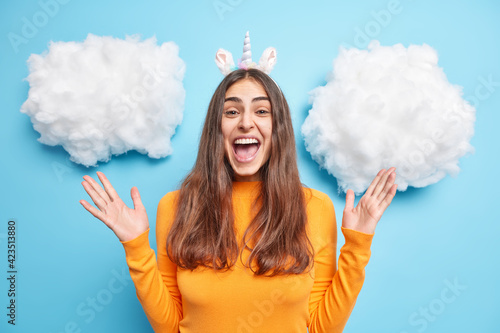 Excited happy brunette woman raises palms feels joyful exclaims loudly reacts on amazing relevation dressed in ornage jumper isolated over blue background white clouds around. Emotions concept