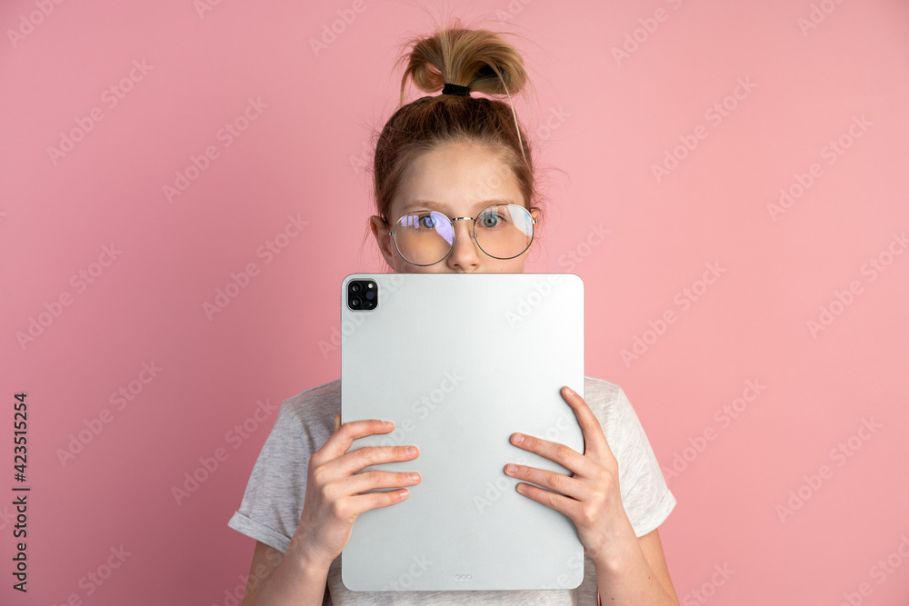 Attractive, blond girl with a tablet and glasses on a pink background. Cute teenager uses a tablet, she covered her face with a tablet, only her eyes are visible. Attractive, blond girl with a tablet and glasses on a pink background. Cute teenager uses a tablet, she covered her face with a tablet, only her eyes are visible.