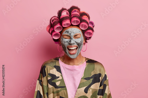Photo of overjoyed ethnic woman laughs out loudly fels very happy enjoys facial treatments wants to have fabulous look makes hairstyle reduces wrinkles with nourishing clay mask. Beauty concept © wayhome.studio 