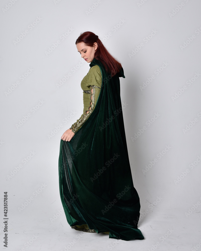 Naklejka premium full length portrait of red haired girl wearing celtic, green medieval gown with fantasy velvet cloak. Standing pose isolated against a studio background.