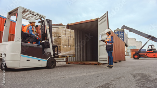 Forklift Driver Loading a Shipping Cargo Container with a Full Pallet with Boxes in Logistics Port Terminal. Latin Female Industrial Supervisor and Safety Inspector with Tablet Managing the Process.
