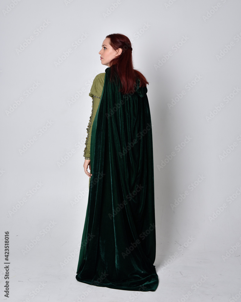 full length portrait of red haired girl wearing celtic, green medieval gown with fantasy velvet cloak. Standing pose isolated against a studio background. full length portrait of red haired girl wearing celtic, green medieval gown with fantasy velvet cloak. Standing pose isolated against a studio background.