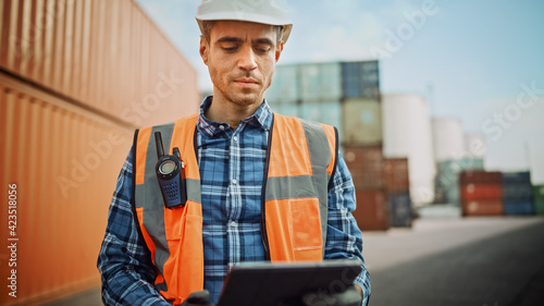 Handsome Caucasian Industrial Engineer in White Hard Hat, Orange High-Visibility Vest, Checkered Shirt and Work Gloves Working on Tablet Computer in Shipping Cargo Container Terminal.