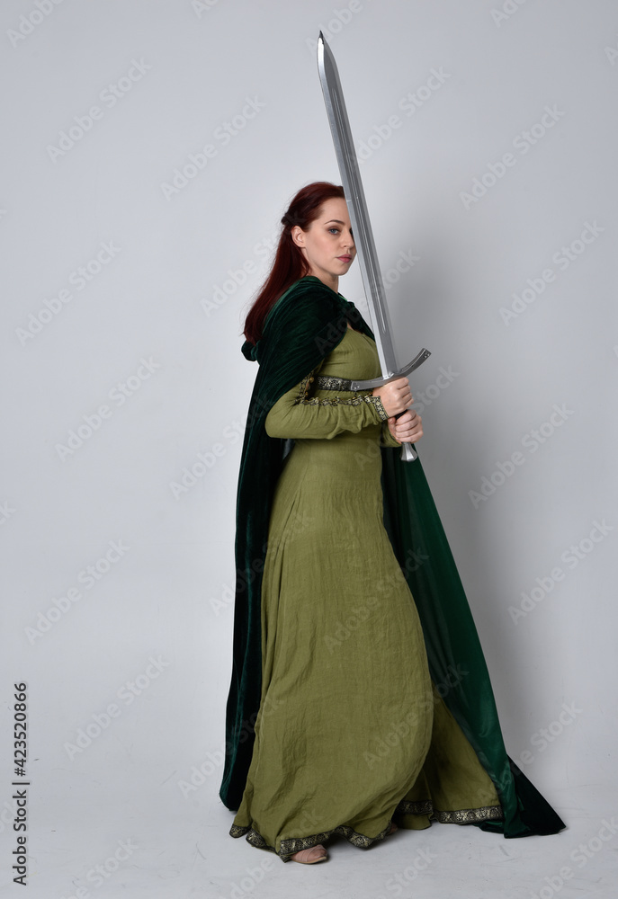 full length portrait of red haired girl wearing celtic, green medieval gown with fantasy velvet cloak. Standing pose isolated against a studio background. full length portrait of red haired girl wearing celtic, green medieval gown with fantasy velvet cloak. Standing pose isolated against a studio background.