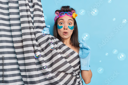 Shocked Asian woman surprised as someone came in bathroom while she was taking shower hides her naked body undergoes beauty treatments makes hairstyle poses indoor soap bubbles around. Hygiene concept