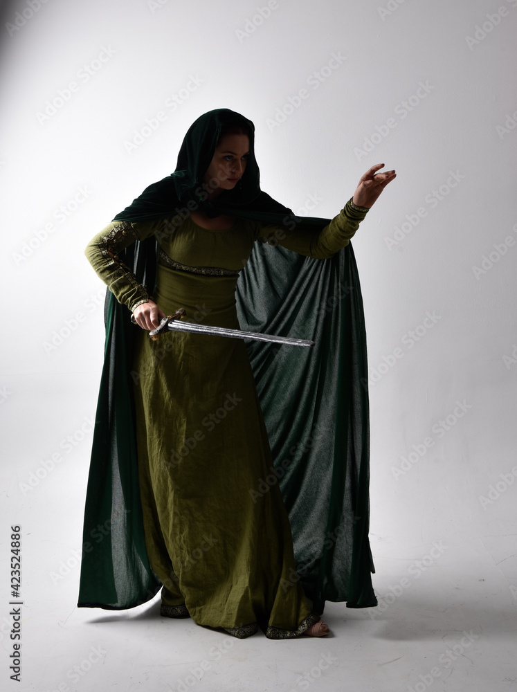 full length portrait of red haired girl wearing celtic, green medieval gown  and velvet cloak with shadowy backlighting. Standing pose isolated against a studio background.
