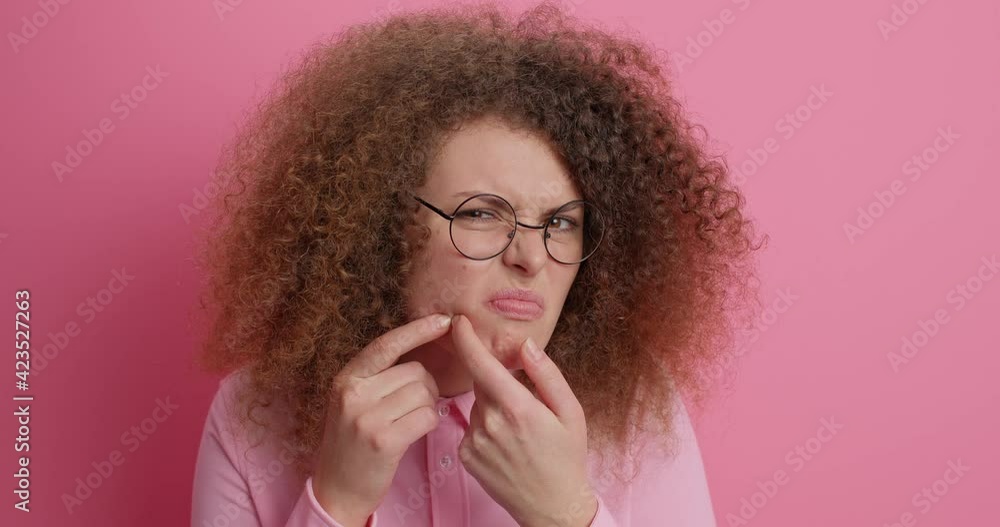 Displeased European teenage girl cannot get rid of acne problem squeezes pimple has puberty period cant deal with blemishes has curly hair wears spectacles and jumper. Problematic skin concept Displeased European teenage girl cannot get rid of acne problem squeezes pimple has puberty period cant deal with blemishes has curly hair wears spectacles and jumper. Problematic skin concept