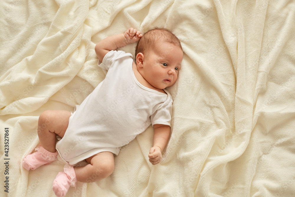 Newborn baby girl or boy lying on blanket on bed looking away, wearing white bodysuit and socks, infant studying world around, has sleepy expression. Newborn baby girl or boy lying on blanket on bed looking away, wearing white bodysuit and socks, infant studying world around, has sleepy expression.