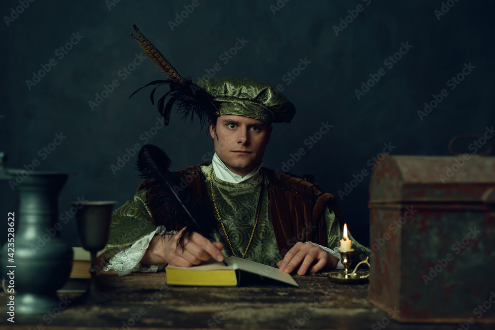 Renaissance man sits behind table writing in a notebook by candlelight in the evening.