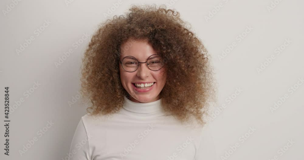 Overjoyed curly young woman appears against white background waves palm excalims hello happy to meet best friend smiles broadly has friendly expression dressed casually wears round spectacles Overjoyed curly young woman appears against white background waves palm excalims hello happy to meet best friend smiles broadly has friendly expression dressed casually wears round spectacles