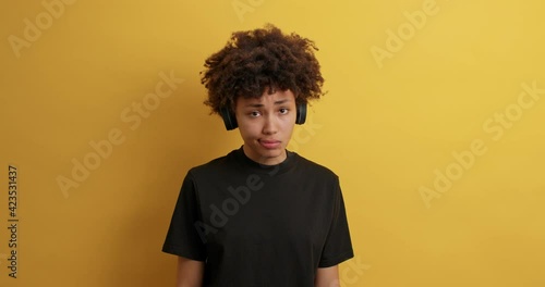 Upset dejected dark skinned woman with curly hair looks sadly at camera being offended by someone listens music to entertain wears wireless headphones black t shirt isolated on yellow studio wall