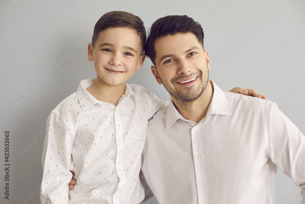 Like father like son. Happy kid and parent. Portrait of cheerful young dad and child in white dress shirts. Little boy and his daddy looking at camera and smiling in studio with light gray background Like father like son. Happy kid and parent. Portrait of cheerful young dad and child in white dress shirts. Little boy and his daddy looking at camera and smiling in studio with light gray background