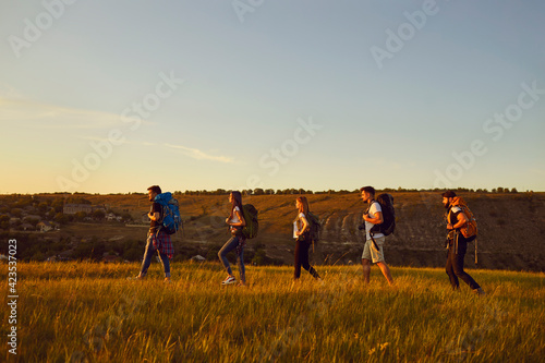 Group of young tourists hikers hiking in row with backpacks in summer field during vacations