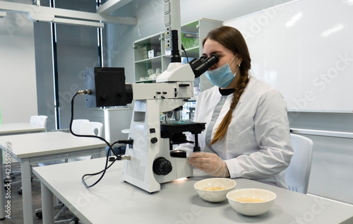 A young woman in a white uniform is working in a laboratory behind a microscope .