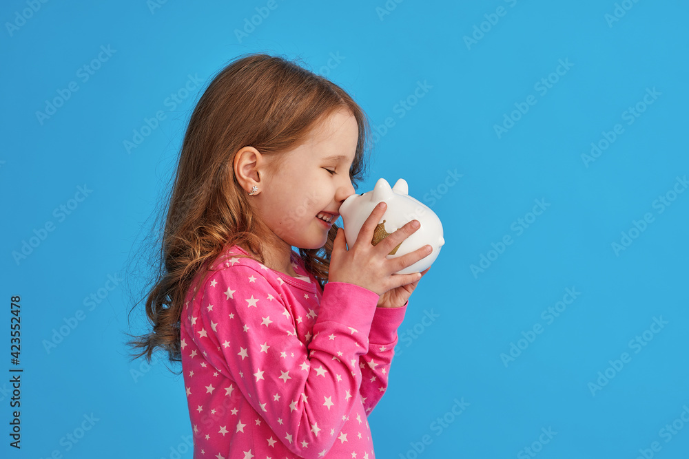 Happy savings. little cute girl with white piggy bank in her hands on blue background in studio. child smiles happily and leans the piggy bank against his nose. concept of saving money for a dream. Happy savings. little cute girl with white piggy bank in her hands on blue background in studio. child smiles happily and leans the piggy bank against his nose. concept of saving money for a dream.