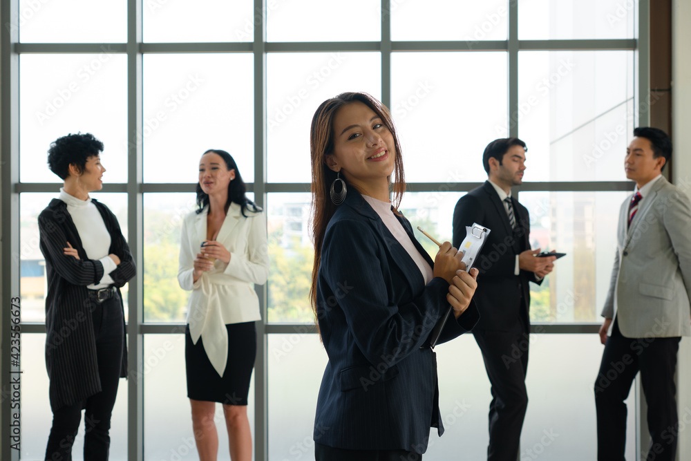 portrait of young asian businesswoman with smiling face stnading in office with background of multiethnic colleagues portrait of young asian businesswoman with smiling face stnading in office with background of multiethnic colleagues