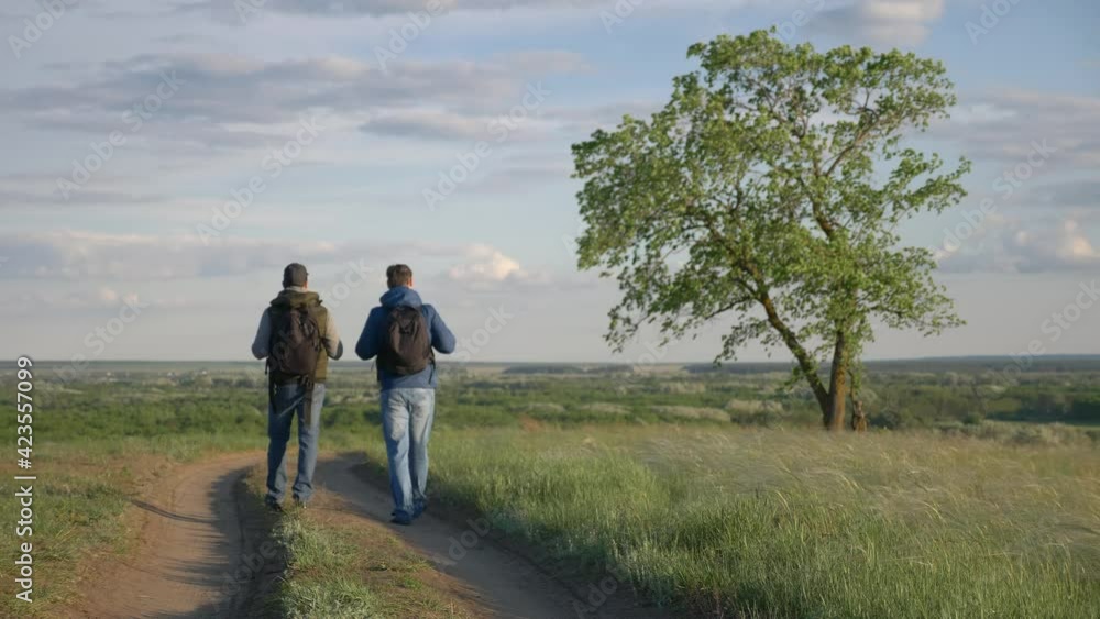 teamwork travel. two man hikers with backpacks walk along trail next to a field with green grass in lifestyle nature. concept travel adventure hiking healthy. two tourists walk walking talking teamwork travel. two man hikers with backpacks walk along trail next to a field with green grass in lifestyle nature. concept travel adventure hiking healthy. two tourists walk walking talking