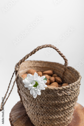 closeup of a wicker basket adorned with a white flower and full of whole almonds on a wooden surface, the background is white and there is space for writing, vertical photo