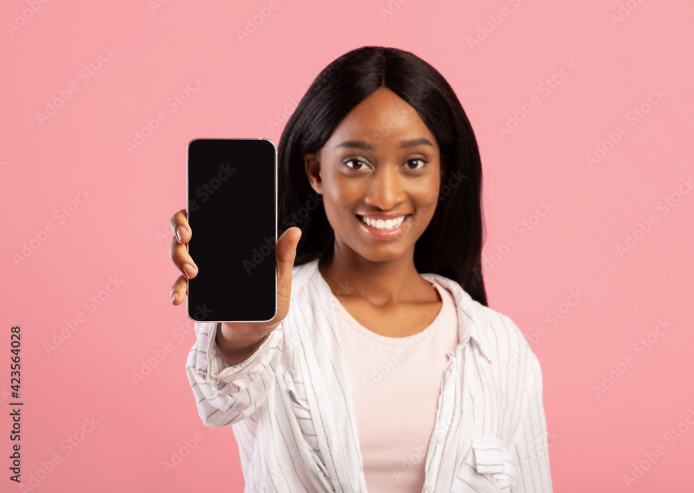 Positive black woman showing cellphone with space for mobile app design on screen over pink studio background Positive black woman showing cellphone with space for mobile app design on screen over pink studio background