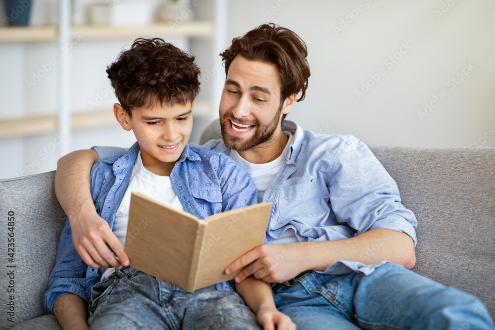Useful time at home. Father and son reading book and smiling, resting together on comfy sofa. Useful time at home. Father and son reading book and smiling, resting together on comfy sofa.