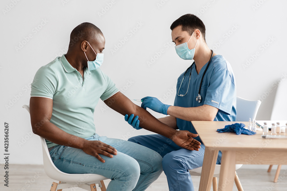Obraz premium Millennial doctor in protective mask and uniform gives shot to african american patient in clinic