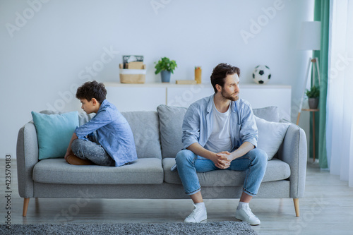 Tela Annoyed father sitting back to back with son on sofa, ignoring each other after