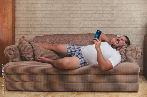 A lazy man yawns while browsing the internet or watching movies on his phone and lying lazily on the couch photo