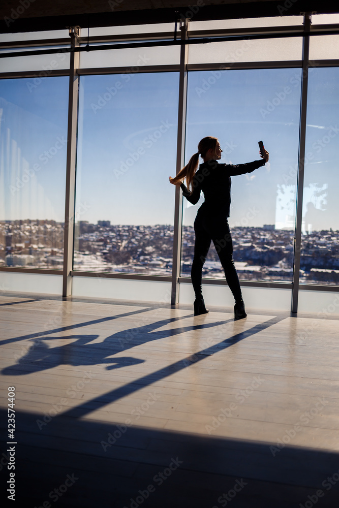 Fototapeta premium A beautiful girl takes a selfie on her phone against the background of panoramic windows in a skyscraper