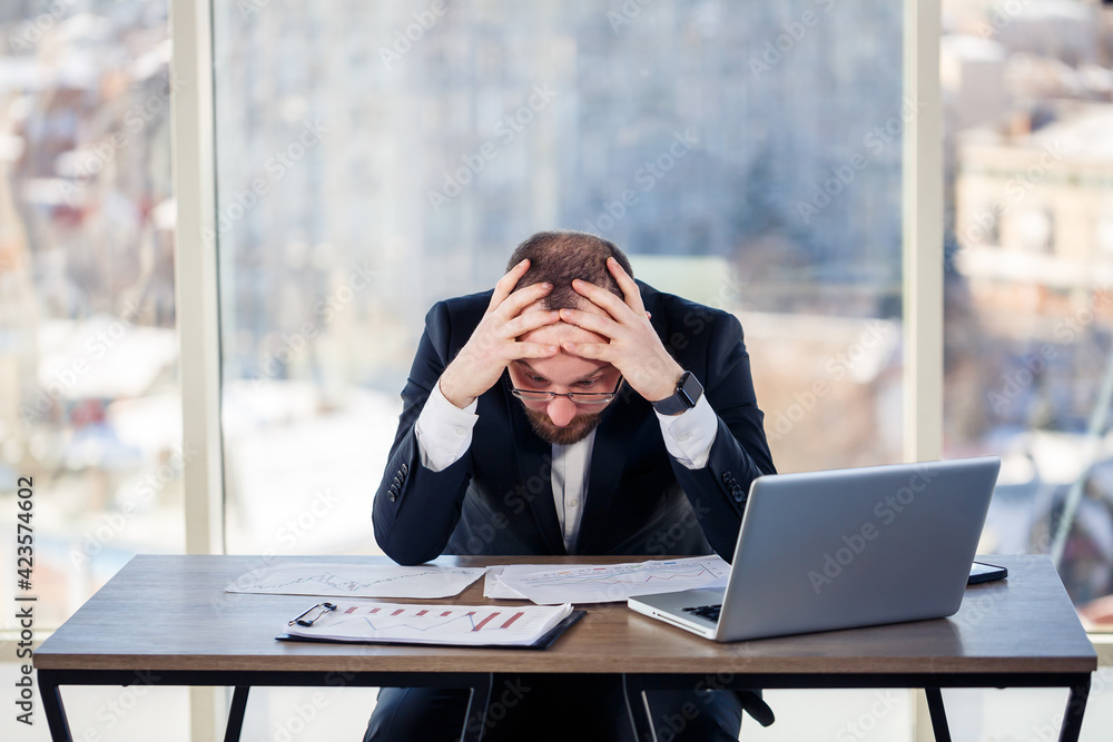 Adult male businessman, teacher, mentor is working on a new project in the office and is nervous. Sits at the table by the large window. He works on a laptop, business problems Adult male businessman, teacher, mentor is working on a new project in the office and is nervous. Sits at the table by the large window. He works on a laptop, business problems