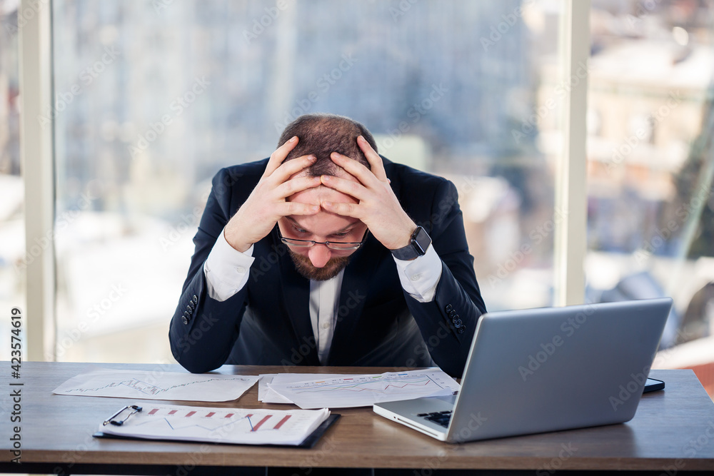 Adult male businessman, teacher, mentor is working on a new project in the office and is nervous. Sits at the table by the large window. He works on a laptop, business problems Adult male businessman, teacher, mentor is working on a new project in the office and is nervous. Sits at the table by the large window. He works on a laptop, business problems