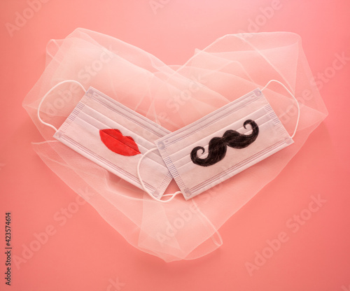 Two disposable protective masks with a mustache and lips painted on them on white heart-shaped veil on pink background. The concept of wedding ceremonies or dates during the coronavirus COVID-19 epide photo