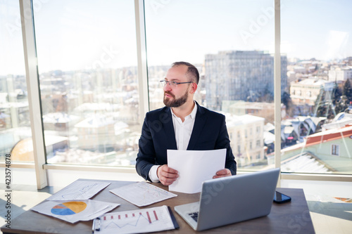 Adult successful male businessman, teacher, mentor is working on a new project. Sits at the table by the large window. It works on a laptop