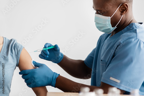Adult african american doctor in protective mask, gloves and uniform vaccinated patient in clinic photo