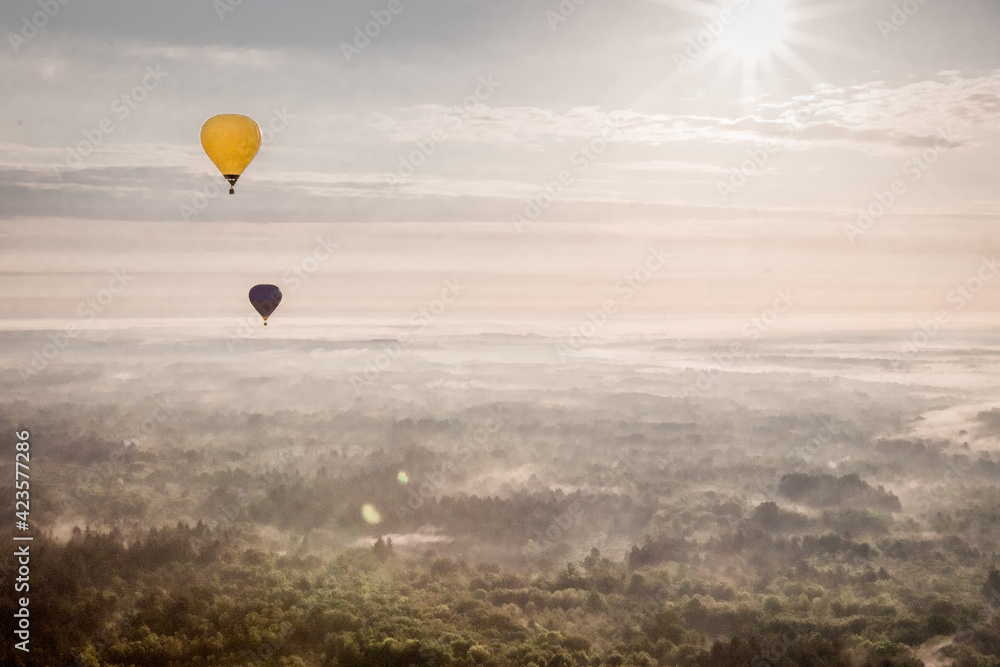ein gelber und ein dunkler Heißluftballon fliegen über einer dunstigen Landschaft in der frühen Morgensonne