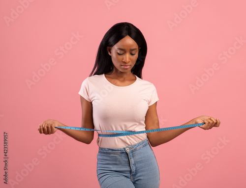 Weight loss. Sad black woman measuring her waist with tape, dissatisfied with body parameters on pink studio background © Prostock-studio