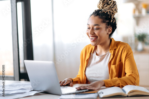 Distance learning. Happy friendly stylish african american female student or freelancer with dreadlocks studying or working remotely from home, chatting by video call uses laptop, smiling