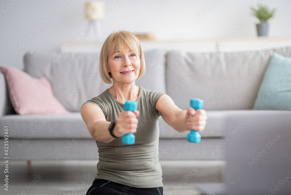 Positive mature woman training with dumbbells near laptop at home, doing online fitness to sports video Positive mature woman training with dumbbells near laptop at home, doing online fitness to sports video