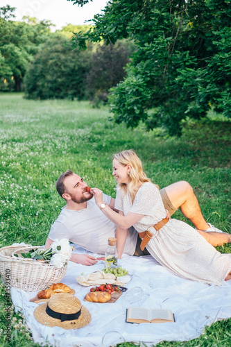 Picnic. Young couple in spring meadow eating tasty food, hugs, smile, love, sitting on grass. Young blond woman in white dress, handsome bearded man, boyfriend. Dating outdoors at summer.