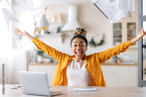 Successful excited happy young african american woman, business lady, manager or freelancer working remotely at home, enjoying career growth or good deal, gesturing with hands, scattering documents