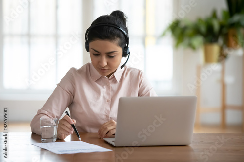 Focused young Indian female employee in headset work online on computer with paper document in office. Concentrated millennial mixed race woman in earphones study distant on laptop at workplace.
