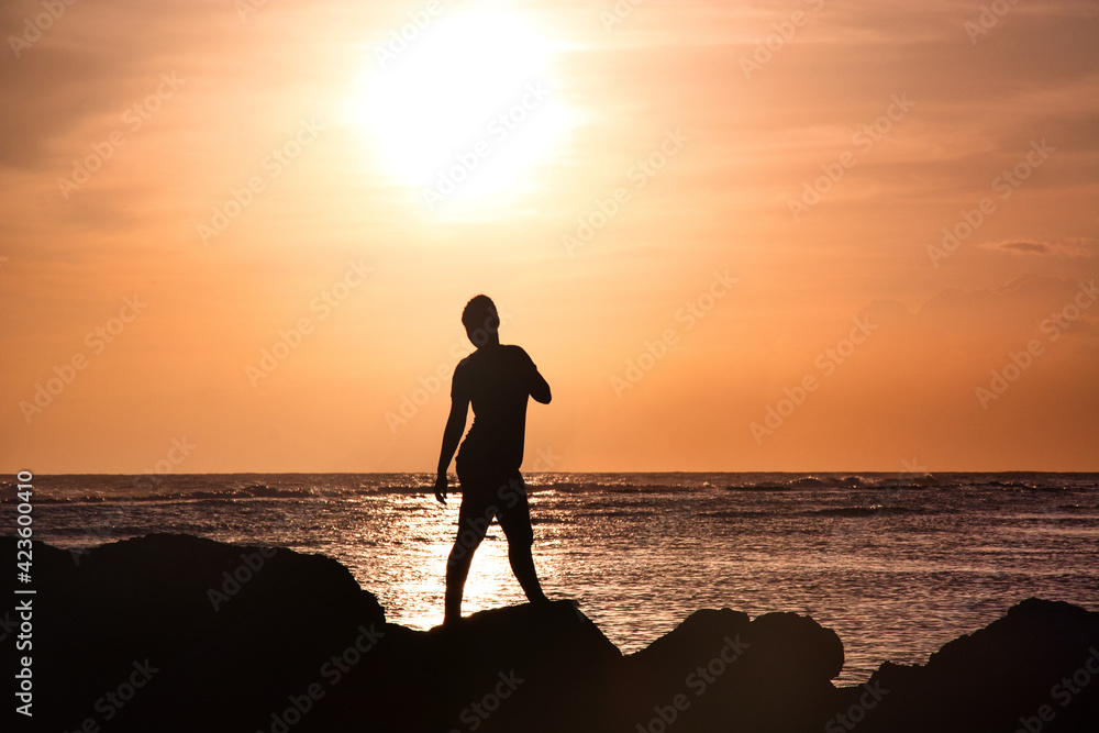 Fototapeta premium Silhouette of woman on beach in orange sunset, standing, looking backwards, relaxed in wide shot