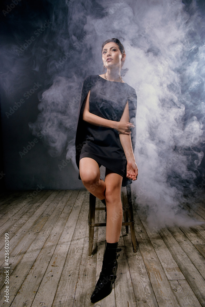 Young elegant woman with perfect stylish make-up and wet brunette hair wearing black tent dress sitting at high chait in dark interior with smoke