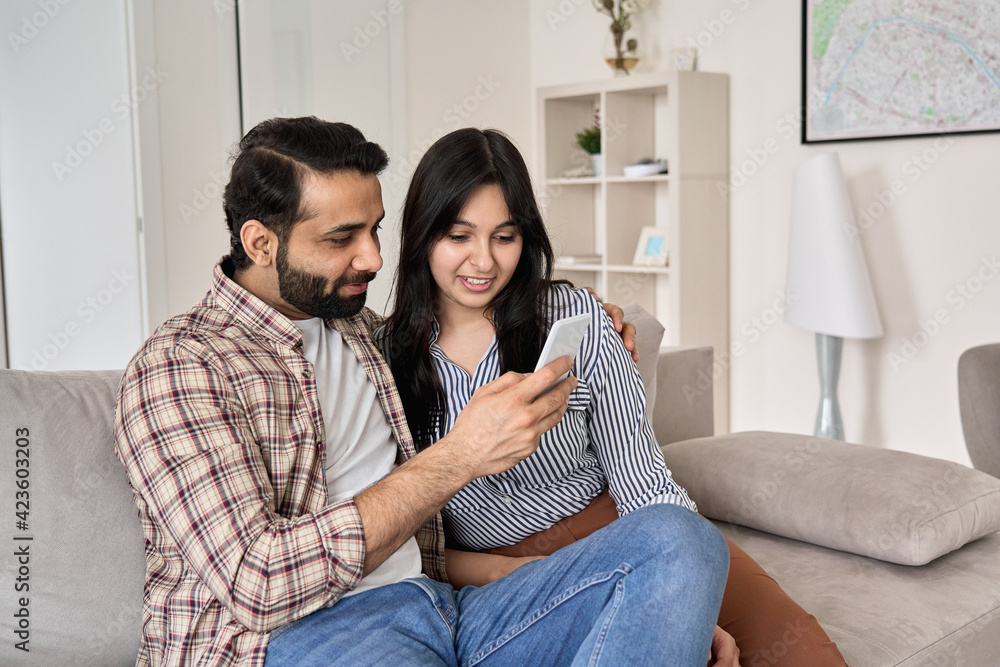 Happy young indian family couple using smart phone looking at cellphone sit on sofa together relaxing at home. Smiling husband and wife checking social media apps or shopping in mobile application. Happy young indian family couple using smart phone looking at cellphone sit on sofa together relaxing at home. Smiling husband and wife checking social media apps or shopping in mobile application.