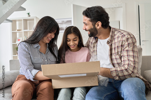 Happy indian family with child daughter unpacking parcel at home. Smiling parents and teen kid daughter opening postal box looking at gift in online shopping delivery package sitting on sofa together.