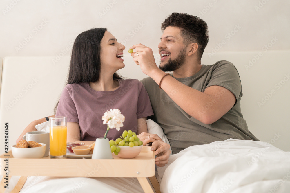 Young cheerful man putting grape in his wife mouth while both sitting under white blanket in double bed and enjoying breakfast