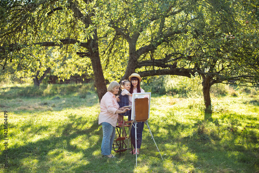 Beautiful summer spring shot of happy three generations family, little kid girl, young mother and mature grandmother, spending time outdoors and painting picture on easel. Summer family leisure. Beautiful summer spring shot of happy three generations family, little kid girl, young mother and mature grandmother, spending time outdoors and painting picture on easel. Summer family leisure.