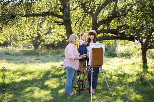 Beautiful summer spring shot of happy three generations family, little kid girl, young mother and mature grandmother, spending time outdoors and painting picture on easel. Summer family leisure. © sofiko14