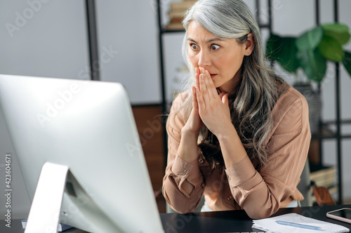 Shocked gray-haired Asian business lady, freelancer, sitting at work desk and receiving unexpected news, message, gesturing her hands while surprised looking at screen, amazed expression on her face