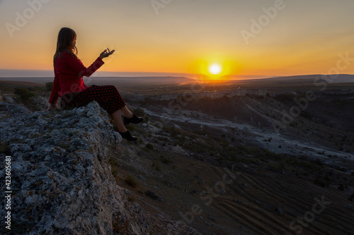Fototapeta Naklejka Na Ścianę i Meble -  A young woman sits on the edge of a cliff, looks at the sun. A beautiful dark-haired girl poses in a dress against the background of mountains. Dawn in the mountains. The concept of unity with nature