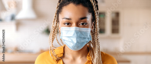 Quarantine precautions. Panoramic headshot african american woman with dreadlocks, in casual wear, stands indoors with a protective medical mask on her face and looks directly into the camera